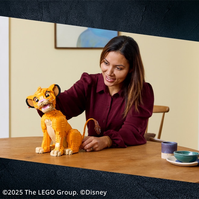Eine Person sitzt an einem Holztisch und baut ein großes LEGO-Modell von Simba aus „Der König der Löwen“ zusammen. Auf dem Tisch stehen außerdem Tassen und Schalen. Im Hintergrund hängt ein Bild an der Wand.